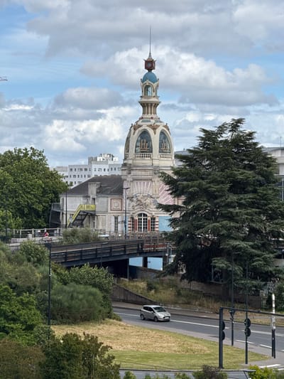 The image depicts the iconic "Maison de l'Erdre" building in Nantes, France, known for its distinctive Art Nouveau architecture and its domed tower topped with a blue spire. The building is situated in an urban environment, with a bridge and a road in the foreground. On the bridge, a person on a bicycle is visible, suggesting movement and activity in the scene. A silver car is driving on the road below, further emphasizing the urban setting. Lush green trees and vegetation surround the structures, creating a contrast between the man-made and natural elements. The sky is partly cloudy, casting shadows and adding depth to the image. The overall scene captures a moment in time within the vibrant city of Nantes, showcasing its architectural heritage and its dynamic urban landscape.