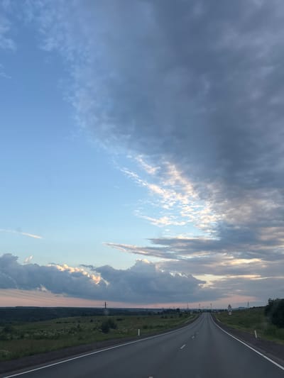 The image depicts a roadside view, likely in or near Бикбарда, Russia, at dusk. The primary subjects are a winding asphalt road and the sky. The road is in the foreground, curving to the right and disappearing into the distance. It is bordered by grassy verges and some scattered vegetation.

The sky is a dominant element, filled with dramatic clouds. The left side of the sky is covered in dark, heavy clouds, while the right side features lighter, wispy clouds illuminated by the setting sun, creating streaks of orange and yellow light. The overall impression of the sky is one of dynamic weather, transitioning from a possibly stormy condition to a calmer, colorful end of the day.

Notable details include a triangular road sign with a number "60" on it, indicating a speed limit, and several utility poles lining the side of the road. The time of day is clearly dusk, indicated by the low angle of the sun and the colored illumination of the clouds. The weather appears to be mixed, with both dark and brightly lit clouds present. The scene evokes a sense of travel or a journey, with the open road stretching towards the horizon. There is no discernible text other than the number on the road sign.
