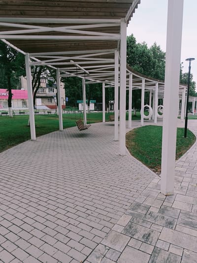 The image depicts a modern outdoor seating area with a covered walkway supported by white columns. The walkway is topped with wooden planks. A paved pathway, made of grey rectangular bricks, runs through the area. Several swings are visible: one wooden swing is hanging under the covered structure, and two circular white swings are suspended further down. Green grass is present on either side of the paved pathway, and trees form a backdrop. In the background, buildings are visible, including one with a pink facade. A street lamp stands on the right side of the frame. The location is identified as the Central Park of Culture and Recreation in Gubkin, Russia. There are no people visible in the image.