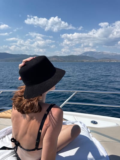 A person is shown from the back, wearing a black hat and a black bikini. They are sitting on a white towel on a boat, with their legs extended. The boat appears to be on the sea, with blue water stretching out to the horizon. In the distance, there are mountains and a clear blue sky with fluffy white clouds. The sun is shining, casting shadows on the boat and the person. The overall scene suggests a leisurely day spent on a boat, enjoying the sea and the sunshine. The person's posture, with their head down and hand on the hat, could imply relaxation or seeking shade from the sun. The clear sky and bright sunlight indicate it is daytime, likely a warm and pleasant day. The presence of mountains in the distance suggests a coastal location.