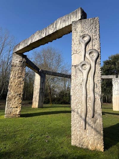 The image displays an outdoor sculptural park located in the city of Mealhada, Portugal, under a clear blue sky. In the foreground, a prominent light-colored stone pillar stands vertically on the right. Its surface is textured, and it features three elongated, spoon-shaped depressions carved into its face, each containing a concentric spiral design at its wider, rounded end. A partial circular relief is visible adjacent to the upper two shapes. This pillar is structurally connected overhead by a thick, horizontal stone beam with a distinctive ridged underside.
To the left, a robust yellowish-brown stone pillar, also with a rough-hewn surface, forms another part of the overarching stone structure. Additional stone pillars, varying in height and design, are dispersed across a vibrant green grassy field in the midground and background. One midground pillar on the left displays dark, abstract stick-figure-like carvings. Long shadows are cast across the grass from the structures, indicating a strong light source. The background is dominated by a dense line of trees, mostly deciduous with bare branches, suggesting a wooded area, with some evergreen trees visible on the far right. No individuals or animals are present in the scene.