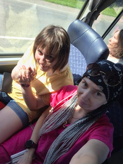 Two women are seated on a bus. The woman in the foreground has braids and is wearing a pink shirt and bandana. The woman in the background is wearing a yellow shirt and denim shorts. The bus appears to be moving, with a glimpse of green grass and pavement visible through the window. The lighting suggests it is daytime.