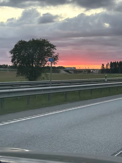 The image depicts a road scene at sunset, viewed from a vehicle. The sky is filled with dramatic clouds, displaying vibrant pink and orange hues typical of a sunset. The setting appears to be a highway, indicated by the paved road with white lane markings and a metal guardrail. Adjacent to the road is a grassy verge and beyond that, some trees and structures are visible.

On the left side of the image, a solitary tree stands prominently, partially silhouetted against the colorful sky. A blue road sign with the white numeral "12" is attached to a pole next to the tree. The sun is low on the horizon, casting a warm glow over the landscape, suggesting it is either early morning or late evening.

The overall mood is serene and picturesque, capturing the beauty of a natural phenomenon against a man-made environment. The focus is on the landscape and the atmospheric conditions. There are no people visible in the frame. The visible text is "12" on the road sign. The location context is Kurna, Estonia.