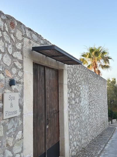 An exterior shot captures a rustic stone wall with a large wooden double door, likely an entrance. Above the door, a dark awning with a textured underside provides shade. To the left of the door, a stone plaque displays the words "Spa & Fitness." A black outdoor light fixture is mounted on the stone wall above the plaque. A palm tree is visible behind the stone wall and to the right of the door, with a clear blue sky overhead. The pathway in front of the entrance is paved with cobblestones. The scene appears to be in the town of Montuïri, Spain, given the architectural style and vegetation. There are no people or animals present.