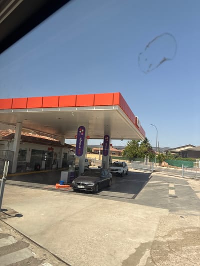 The image shows a view from inside a vehicle looking out at a gas station in Villa Del Prado, Spain. A red-and-white awning covers several gas pumps. Two cars are visible at the pumps: a dark gray sedan in the foreground and a white SUV behind it. A building with a tiled roof is situated behind the pumps. In the background, houses and trees are visible under a clear blue sky. The scene appears to be set on a sunny day.