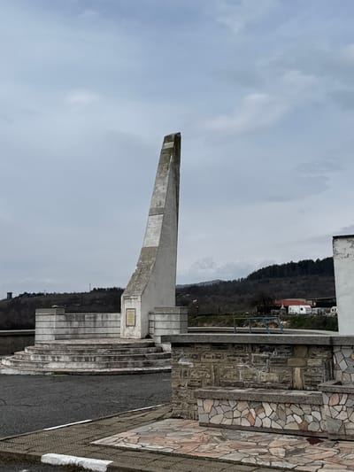 An outdoor scene in Баня, Bulgaria, is depicted under an overcast, light grey sky. The central subject is a tall, angular concrete monument, off-white in color and showing weathering, with a pointed apex leaning towards the viewer's left. It stands on a multi-tiered circular concrete platform featuring steps and flanking concrete walls. A rectangular plaque, appearing to be brass or bronze, is affixed to the front base of the monument. To the immediate right of the foreground, a low wall constructed of irregular grey and brown stones is visible, topped by a flat concrete slab. Paving stones of various irregular shapes and colors (pink, beige, grey) are laid out in front of this wall. In the midground, behind the monument, a Bulgarian flag (white, green, red stripes) is partially visible. Further back, on a distant hill to the left, a circular stone tower is present. To the right of the monument, several vehicles, including two white recreational vehicles (RVs) and a smaller white vehicle, are parked near a small white building with a dark roof. The background consists of rolling hills densely covered with bare deciduous trees or dormant vegetation. No individuals or active interactions are present in the image.