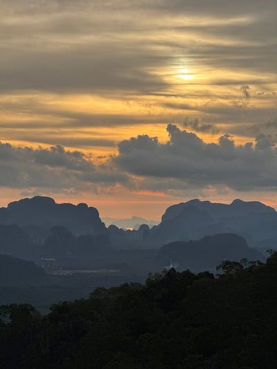A wide-angle, low-angle shot captures a dramatic sunset over a mountainous landscape in Ban Khao Tang, Thailand. The sky is dominated by streaks of orange and yellow, with the sun a bright, hazy disc peeking through scattered clouds. Below the sky, a series of dark, silhouetted mountain ranges recede into the distance, their jagged peaks creating a stark contrast against the vibrant sky. The foreground is filled with the dark green silhouettes of trees and dense foliage, suggesting a forested area or a viewpoint overlooking a natural expanse. The overall impression is one of natural beauty and dramatic lighting.