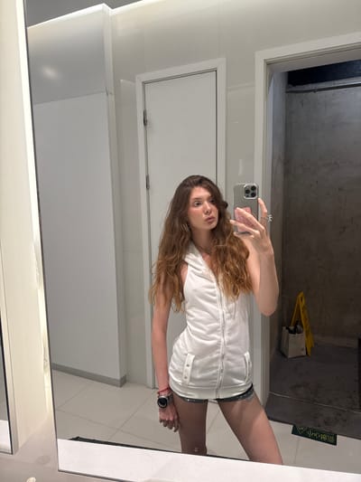 A woman with long brown hair takes a selfie in a bathroom mirror. She wears a white sleeveless top and denim shorts. She holds a smartphone in her left hand, showing its back with a pink accessory, and wears a watch on her right wrist. The bathroom features white walls and a door, a gray floor with a yellow and black caution sign, and a darker gray area in the upper background. This photo was taken at the location of the bathroom, situated in Hongkou District, China.