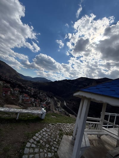 The image depicts a landscape in the city of Bov, Bulgaria, with a view from an elevated position. The foreground features a portion of a white wooden structure, possibly a gazebo or viewpoint shelter, with a blue roof. To the right of this structure, a stone-paved path and grassy area are visible. Below and slightly to the left, a village or town nestled in a valley can be seen, with houses and buildings clustered together, many featuring red rooftops. A mountain rises sharply behind the town, casting a significant shadow over the area. The sky above the mountain is bright blue, filled with scattered white clouds. The sun is shining through some of the clouds, creating a diffused, bright light. A wooden bench is visible near the stone-paved path. The photograph appears to have been taken during the day, with a clear sky and good visibility.