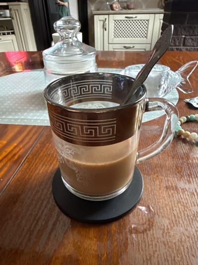 The image, taken indoors in Krapivka, Russia, displays a glass mug containing a light brown, creamy beverage, likely coffee, resting on a black circular coaster. A silver spoon is placed within the mug. The mug features a prominent white Greek key pattern as a decorative band around its upper section, complemented by white swirly designs further down. The mug and coaster are positioned on a glossy wooden table, which exhibits a clear grain pattern.
In the background, partially visible to the left, is a light-colored kitchen cabinet or piece of furniture with a lattice-patterned door and silver handles. A dark, textured wall or surface is also seen in the far background. To the right of the central mug, a clear glass decorative object, possibly resembling a fish, and a beaded bracelet with white, beige, and light blue/green beads are present. Other clear glass items, including a small jar with a decorative stopper and a textured glass dish or holder, contribute to the table arrangement.
The scene suggests a casual setting, possibly a kitchen or dining area, with a beverage prepared for consumption. The lighting indicates a daytime shot, though no specific time of day or weather is discernible. No visible text is present in the image, only decorative patterns.