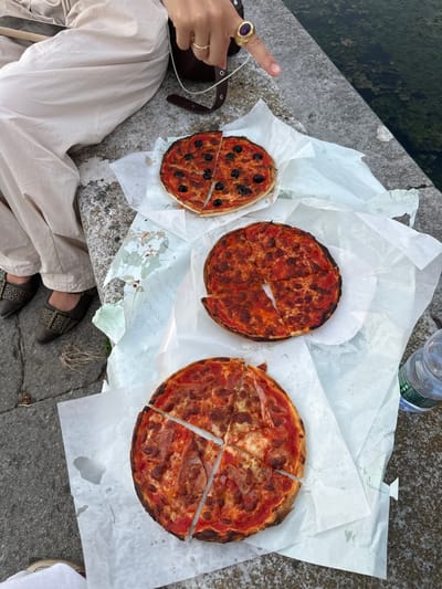 Three individual pizzas, likely personal size, are arranged on white paper on a grey stone ledge. The uppermost pizza is topped with black olives and sliced. The middle pizza appears to be a plain cheese and tomato pizza, also sliced. The bottom pizza is topped with cheese and what appears to be cured meat, and is sliced. A person's lower legs and feet are visible on the left, wearing light-colored pants and dark, buckled shoes. A hand with a large purple stone ring is visible in the upper right, pointing towards the pizzas. A plastic water bottle is also visible to the right of the pizzas. The setting appears to be outdoors, near a body of water, possibly a canal or river, with some vegetation visible in the water. The architecture of the surrounding area, typical of Padua, suggests a location along the Navigli canal system or near the Prato della Valle.