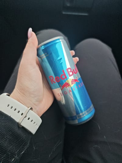 The image shows a person holding a can of Red Bull Sugarfree. The can is primarily blue with red lettering. The text "Red Bull" and "SUGARFREE" are clearly visible on the can. The person's hand, with painted nails and a white smartwatch, is holding the can. The background is dark, suggesting an indoor setting or perhaps a car interior. The overall scene appears casual, with the person likely consuming the beverage. No specific time of day, weather, or strong emotions are discernible from the image. There are some faint Russian characters visible on the can, which are difficult to read fully. Based on the provided location context, it is possible the can is intended for sale in or was purchased in Shuya, Russia.