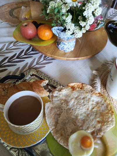 A high-angle, close-up shot shows a table setting with a breakfast spread, likely in Alexandrovskaya, Russia. In the foreground, a cup of coffee sits on a yellow patterned saucer. Beside it, a flatbread, possibly chud, is visible along with a small, hard-boiled egg cut in half, revealing its yolk. To the left, a silver tray holds pieces of pastry or bread. Further back, a plate contains an apple and an orange, and a bouquet of white flowers in a patterned vase rests on a wooden tray. The table is covered with a white cloth featuring a brown leaf pattern. The overall atmosphere is of a tranquil morning meal.