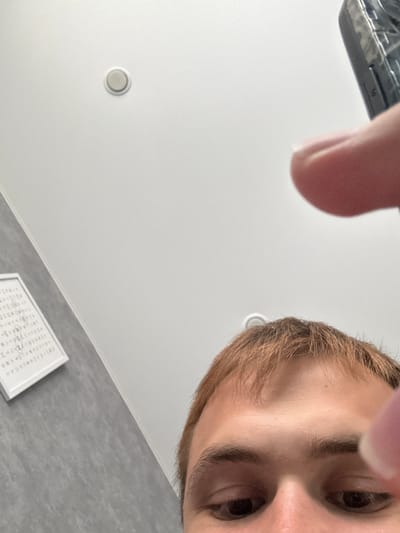 The image is a close-up, low-angle shot of a person's face, specifically their eyes, nose, and forehead, looking upwards. The person has light brown hair and appears to be a young individual. They are indoors, as indicated by the white ceiling with a circular fixture and a grey, textured wall in the background. In the bottom right corner, a framed piece of white paper with text is visible. The lighting suggests it is daytime. There is no discernible activity or event, and the emotion is neutral. The location is Tlustenkhabl, Russia.