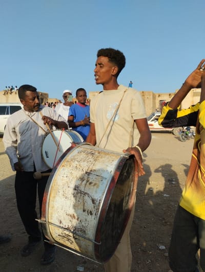 A group of men play drums outdoors on a sunny day. The man in the foreground holds a large, well-worn drum. Another man, wearing a button-down shirt, beats a smaller drum alongside him. A younger man in a blue shirt stands nearby, observing. Other people, cars, and buildings are visible in the background. The scene appears lively with musical performance in a public space.