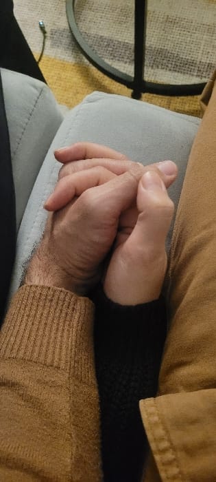 A close-up shows two hands clasped together, resting on a light gray cushion. The bottom hand is covered by a brown, ribbed sweater sleeve. The top hand appears lighter in complexion. The person is wearing a brown jacket or shirt. A corner of a black table leg and a portion of a striped rug with yellow, gray, and black are visible on the left side. The lighting is soft, highlighting the textures of the clothing and the wrinkles on the hands.