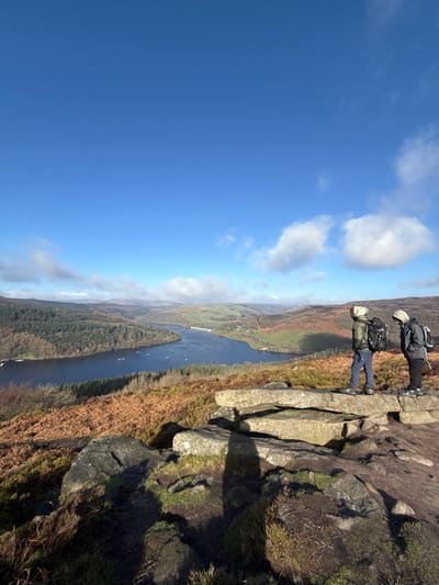 The image captures an expansive outdoor scene on a clear, bright day under a blue sky with scattered white clouds, located in the Peak District, High Peak, United Kingdom, overlooking Ladybower Reservoir. In the foreground, two individuals, dressed in hiking attire and carrying backpacks, stand on a flat, light-grey rock outcrop amidst brown heather and green moss. The individual on the left wears a khaki jacket, light blue trousers, and a light-colored hood, while the individual on the right wears a dark hooded jacket and dark trousers. Both are facing right, observing the distant landscape. Below them, the dark blue waters of Ladybower Reservoir stretch across the mid-ground, containing several small boats and spanned by a distant grey arched viaduct. The reservoir is flanked by dense green coniferous forest on the left bank and rolling hills with mixed brown and green vegetation on the right. Further rolling hills extend into the background beneath the horizon.