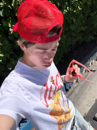 A person wearing a red mesh trucker hat and a white T-shirt with a yellow and red graphic is holding pink sunglasses outdoors. The person is also wearing a gold necklace with a clover pendant and several colorful beaded bracelets. The background shows green foliage. The setting appears to be an outdoor location.
The exact location in Indian Wells, United States of America, is not discernible from the image.