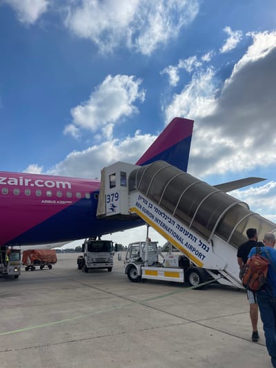 A pink and blue Wizz Air Airbus A321neo airplane is parked on an airport tarmac under a cloudy blue sky. A mobile passenger staircase is connected to the aircraft's door, with the words "BEN GURION INTERNATIONAL AIRPORT" printed on its side in English and Hebrew. Two men, one with a red backpack, are walking up the stairs, presumably boarding the plane. Several ground vehicles, including a baggage tug and a tow truck, are visible near the aircraft. The scene appears to be at Ben Gurion International Airport near Tel Aviv, Israel, not the city of Yagel.
