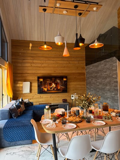 The image shows an interior scene, likely a living room or dining area in Марусино, Russia. A wooden wall is adorned with a large picture of a fireplace and a white wall-mounted unit. A modern light fixture with multiple pendant lamps hangs from the ceiling. A long wooden table is set with various dishes, drinks, and decorative items. White chairs surround the table. A blue sofa is situated near the table, decorated with pillows. The floor is covered with marble tiles.