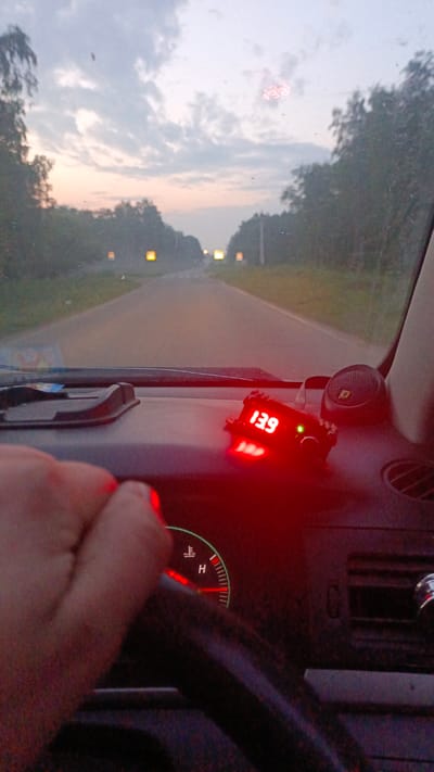 The image shows the interior of a vehicle, looking forward through the windshield at a road at dusk. A hand is visible on the steering wheel. On the dashboard, a red digital display shows "13.9", and a thermometer gauge indicates a temperature between "H" and a lower mark. The road is flanked by trees, and several illuminated signs or lights are visible in the distance. The sky is overcast with a soft, diffused light. The image is grainy and appears to have been taken with a low-quality camera or in low light conditions. The location is identified as Zuy, Russia.