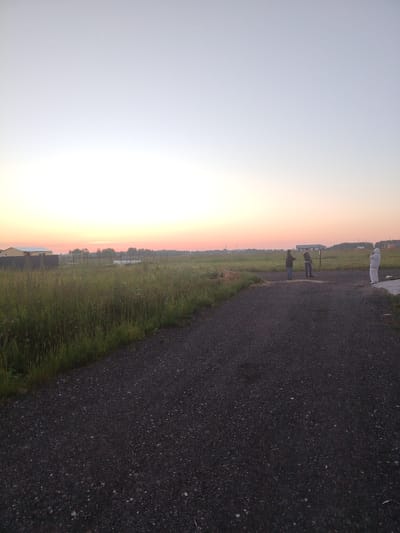 In Rastunovo, Russia, at dusk, a gravel road winds through a grassy field with a building and a fence in the background. Three individuals are present. Two men stand on the gravel road, one facing left and the other facing forward. To the right, a third person, possibly a woman, is wearing a white protective suit and is facing the viewer. The sky exhibits a gradient of pale blue to soft pink, indicating the setting sun.