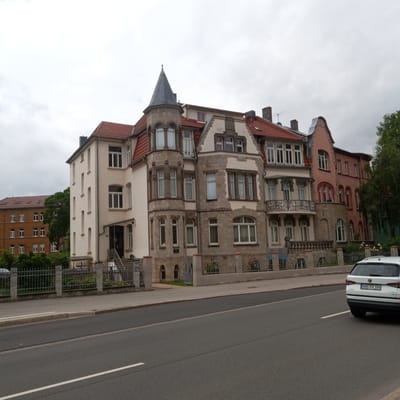 This is a photograph of a street scene in Eisenach, Germany, featuring several apartment buildings. The main building in the foreground is a multi-story structure with a red tiled roof, stone accents, and a prominent turret. It has numerous windows of various sizes and shapes. The building to its left is a lighter color with a similar roofline and many windows. Further to the left, another apartment building with a brown facade and a mansard roof is visible. To the right of the main building, a pink-colored apartment building is partially visible, also with a red tiled roof. A white SUV is parked on the right side of the street, facing away from the camera, with its license plate visible. The street itself is paved, with a sidewalk and a low decorative fence in front of the buildings. The sky is overcast. The overall impression is that of a residential street with older European architecture.