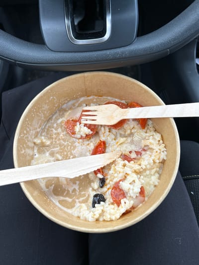 The image shows a partially eaten bowl of risotto in a brown cardboard container. There are wooden cutlery items, a fork and a knife, stuck in the food. The risotto appears to contain rice, tomatoes, olives, and cheese. The bowl is resting on black fabric, possibly clothing, inside of a car, with the steering wheel visible to the left. The location is in a car somewhere in Valence, France.