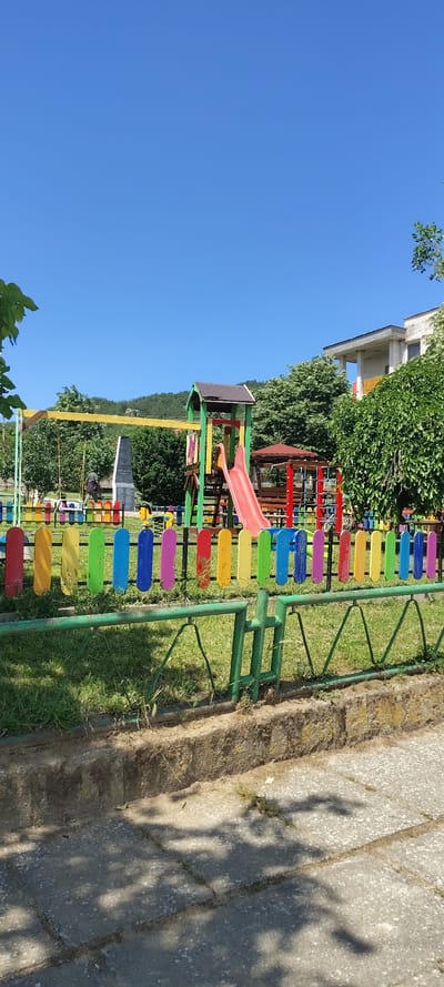 The image shows a playground in Beguntsi, Bulgaria. The playground equipment includes a slide and swings, and is surrounded by a colorful fence. The playground is next to a concrete pathway and a grassy area. The sky is blue. There are trees and a building in the background.
