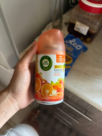 In Dromolaxia - Meneou Municipality, Cyprus, a person's hand is holding an Air Wick Essential Oil air freshener can. The can has an orange cap and a label featuring oranges and flowers. The label text indicates "FRAGRANCE WITH NATURAL Essential Oil" and "Anti Tabac Anti Tobacco." Behind the can, a bag of "oleo tics" snacks and a large jar with a red lid filled with a dark liquid are visible on a wooden surface. The background shows a textured wall and what appears to be kitchen cabinetry.