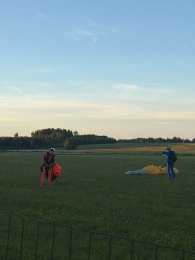The image depicts an outdoor scene with a wide expanse of green grass, likely a field or airfield, under a pale blue sky with a few wispy clouds. The horizon is marked by a dense line of trees. In the foreground, a dark metal fence runs across the bottom of the frame.

Two individuals are present, both appearing to be skydivers. On the left, a person in an orange jumpsuit is standing, facing away from the camera and towards the right. They have a red parachute bag attached to their back and appear to be adjusting it. On the right, a person in a blue jumpsuit is also standing, facing left and slightly away from the camera. This individual is also wearing a black backpack and is looking down at a yellow and gray parachute lying on the ground in front of them. The skydiver on the right appears to be gathering or preparing the parachute. The lighting suggests it is either early morning or late afternoon.

The scene is devoid of any urban structures or landmarks that would specifically place it within the city of Limbaži, Latvia. The environment appears to be rural, consistent with an area used for outdoor activities like skydiving, which often takes place at airfields located outside of city centers.
