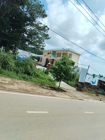 The image shows a street in Pờ Y, Vietnam. A two-story building with a red cross is set back from the road behind a patch of green vegetation. The building has a light-colored facade and a green roof. Signs are visible to the left and right of the building. Trees are present on the left side of the frame, and power lines run across the top of the image. A road with painted white lines is in the foreground. More vegetation and signage are visible on the right side of the frame. The sky is cloudy.