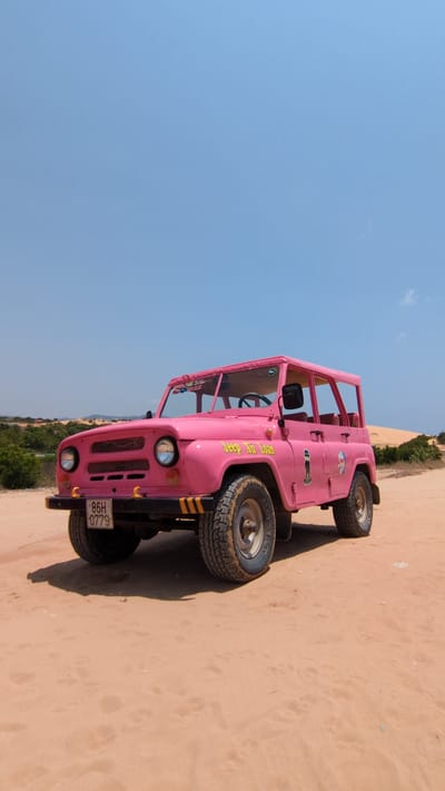 A pink, roofless off-road vehicle is parked on a sand dune in Phuoc Dinh, Vietnam. The vehicle has visible license plate "86H 0779" and writing on its side in yellow font. The location is sandy with sparse vegetation in the distance. The sky is clear and blue.