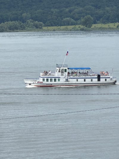 A white passenger boat with a blue canopy is sailing on a body of water. The boat has multiple decks and appears to be carrying several passengers who are visible on the upper deck. A Russian flag is flying from a pole at the front of the boat. In the background, there is a lush green forested hillside. A thin dark line, possibly a wire, stretches horizontally across the foreground. The scene is set on a river, possibly the Volga River, near the city of Sergeevka, Russia. The overall impression is that of a calm, scenic day on the water.