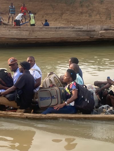 A group of people are crowded onto a wooden boat. They are carrying various luggage items like backpacks, suitcases, and bags. The boat is on a body of water. On the left of the boat, people appear to be on the shore. The picture is taken at an angle.