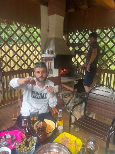 In the city of Soloslovo, Russia, a man with a beard and sunglasses is seated in a wooden chair on a wooden deck, wearing a white sweatshirt with "ELEGANCE" printed on it. He is gesturing with his left hand and holding a black object in his right hand. To his right, a dog with brown and grey fur is lying down. Behind them, a brick barbecue with a fire is visible, and another man in a dark shirt with a flamingo pattern is standing and appearing to tend to the fire. The environment is an outdoor gazebo with wooden lattice walls, surrounded by greenery. On the table in front of the seated man are various items including bottles of alcohol, a glass of amber liquid, a bowl of olives, and a pink tray with sliced fish. A yellow serving tray with what appears to be shrimp shells is also present. The scene depicts a relaxed outdoor gathering.