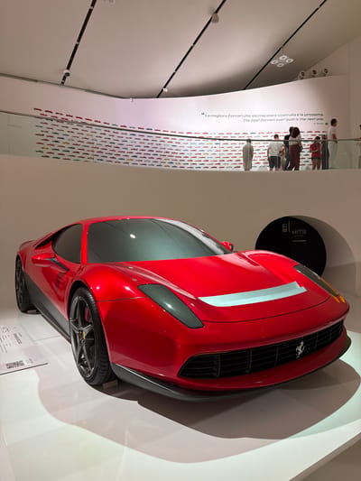 The image shows a vibrant red Ferrari sports car on display in a museum. The car is positioned centrally in the frame, viewed from the front and slightly to the left. It features a sleek design with sharp lines and a metallic finish. The interior appears to be tinted, obscuring the driver's cabin.
In the background, a white curved wall displays a collage of small Ferrari car images in various colors, arranged in rows. Above this display, a quote in both Italian and English is visible, attributed to Enzo Ferrari: "La migliore Ferrari che sia mai stata costruita è la prossima. The best Ferrari ever built is the next one."
A group of people, including men, women, and children, are standing behind a railing, observing the exhibit. They are casually dressed and appear to be visitors. Some are looking at the car, while others are engaged in conversation or looking at the display.
To the right of the car, a black circular structure with the word "HITS" and "SALA JUKEBOX" is visible, possibly an interactive display or part of the museum's audio-visual setup.
In the lower left corner, a white placard with QR codes and text, likely providing information about the car or the exhibition, is partially visible. The floor is a reflective white surface, casting shadows from the car and the surrounding elements.
The overall impression is of a modern museum environment dedicated to the Ferrari brand, showcasing their iconic vehicles and heritage. The lighting is bright and focused, illuminating the car and the exhibits. This scene is consistent with the Ferrari Museum in Modena, Italy, which features such displays celebrating the history and innovation of the brand.
