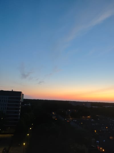 This is a high-angle, full-frame, horizontal photograph capturing a twilight sky over the city of Tilburg, Netherlands. The sky transitions from a clear, light blue at the top to a soft gradient of orange and yellow near the horizon, indicative of sunset or sunrise. A thin, wispy cloud is visible in the upper portion of the sky. In the foreground, a dark silhouette of a treeline stretches across the middle ground, separating the sky from the urban landscape below. To the left, a tall, modern building with a dark facade and visible window patterns stands out. The lower part of the image reveals a dimly lit urban area, with scattered lights from street lamps and buildings visible amongst the dark shapes of trees and structures. The overall impression is a serene, twilight cityscape.