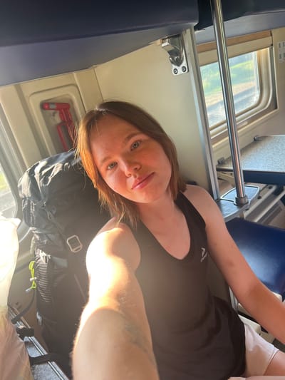 A young person with shoulder-length brown hair and freckles is taking a selfie on a train. They are wearing a black tank top and white shorts. A black backpack is to their left, and a white plastic bag is to their far left. The train car has blue seats and a window showing greenery outside. A red emergency handle is mounted on the wall. The image appears to be taken in Pugachevo, Russia, as indicated by the presence of Russian text on signs and train car features.