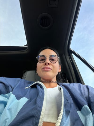 A woman is taking a selfie inside a vehicle. She is wearing a white ribbed tank top, a blue and light blue color-blocked windbreaker, round glasses, and a black choker necklace. Her face is centered in the frame, with her head tilted slightly. The sunroof of the vehicle is open, revealing a cloudy sky. The interior of the car is dark, with a visible air vent on the ceiling. The setting is the city of Sholokovo, Russia.
