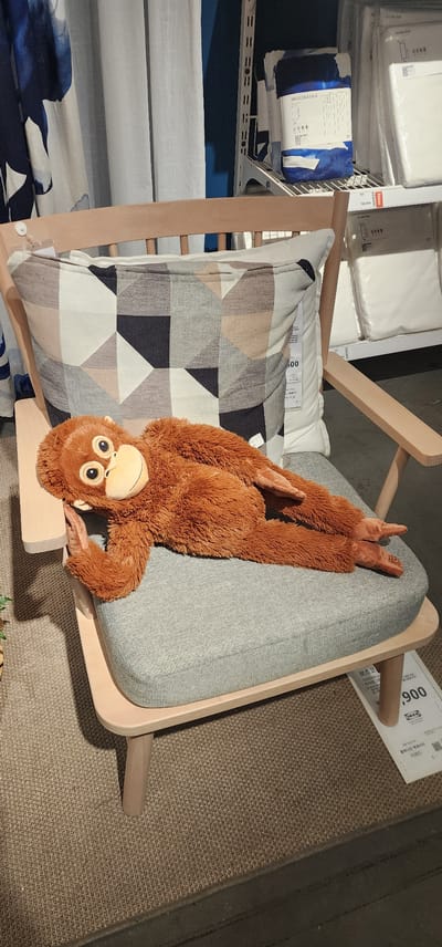 A plush orangutan toy is lying on its back on a light gray upholstered chair, with its arms and legs spread out. The chair has a light-colored wooden frame and is placed on a textured brown rug. To the left of the chair, there are shelves displaying various items, including what appear to be bedding products with blue and white patterns. A white pillow with a geometric pattern of gray, black, and beige shapes is visible behind the orangutan. A white tag with Korean text and the number "900" is attached to the chair. The scene appears to be indoors, likely in a retail store such as IKEA, given the visible IKEA logo on the tag. The lighting suggests it is daytime.
