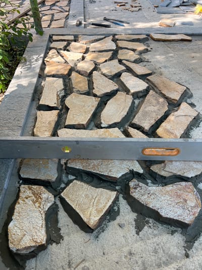The image captures a section of landscaping work in progress in Menenkyovo, Bulgaria. The foreground shows a pathway being laid with irregular, flat stones set in mortar. A spirit level is positioned horizontally across the laid stones, indicating an effort to ensure a level surface. The stones are predominantly light brown and beige, with some showing hints of reddish-brown and white speckling. The mortar is dark gray and fills the gaps between the stones. In the upper portion of the image, more stones are visible, both laid and stacked, along with some gardening tools. To the left, lush green foliage suggests an outdoor setting, possibly a garden or park. The overall scene depicts manual construction and meticulous attention to detail in creating an outdoor pathway.