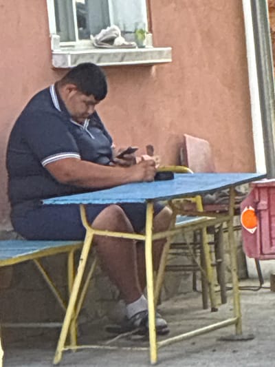 The image depicts an outdoor scene in Radievo, Bulgaria, featuring a single adult male seated at a casual table and bench arrangement. The man, with dark, short hair and a medium complexion, is wearing a navy blue polo shirt with white and blue striped sleeves, navy blue shorts, white socks, and dark shoes. He is positioned on a blue bench, leaning forward with his head tilted downwards, intently focused on a dark-colored smartphone held in both hands. An additional dark, rectangular object, possibly a wallet or another device, rests on the blue tabletop to his left. The table and bench both feature yellow metal frames. In the background, a salmon-pink stucco wall is visible, with a white-framed window on which a light-colored, crumpled object and a small green plant in a pot are placed. To the far right, a vertical grey and white pipe or column stands, alongside a reddish-pink crate adorned with an orange circular sticker. The ground is a light grey, possibly concrete.