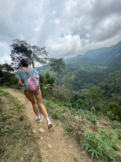 A woman stands on a dirt path in Suối Giàng, Vietnam, facing a forested valley under a cloudy sky. She wears a light blue shirt, shorts, lavender socks, and sneakers. A floral backpack is slung over her shoulder. The path is flanked by vegetation, and a steep drop-off is visible. The landscape is lush and green, and the sky is overcast.