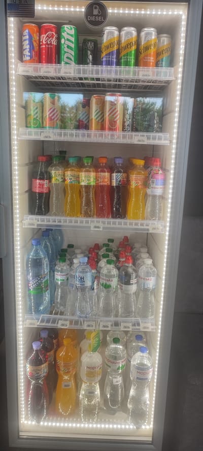 This is a vertical, eye-level, full shot of a brightly lit refrigerated display case filled with various beverages. The display case appears to be in a store or vending area. The top shelf holds several cans of soda, including Fanta, Coca-Cola, Sprite, and Schweppes in different flavors like blueberry, classic mojito, tangerine, and bitter lemon. Below the cans, several bottles of juice and lemonade are arranged, with labels in a Cyrillic script. The middle shelves are stocked with numerous bottles of water, mostly clear plastic bottles with various labels, some featuring green and blue accents. The bottom shelves also contain more bottled drinks, including some with darker liquids and brightly colored caps. The overall impression is one of a well-stocked beverage cooler, with a focus on refreshing drinks. The lighting is artificial and prominent, highlighting the products. Text visible on the display includes brand names like Fanta, Coca-Cola, Sprite, Schweppes, and what appears to be a Ukrainian brand "Моршинська" (Morshynska) on many of the water bottles. There is also a "DIESEL" logo visible at the top. The overall setting is clean and organized.