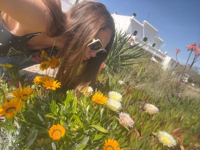 A woman in sunglasses stands in front of a mix of yellow and white flowers, with a white building in the background under a clear blue sky. The location is likely in La Marina De Creixell, Spain.