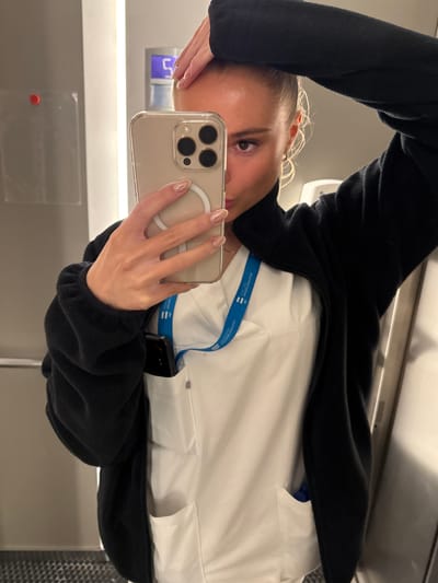 A woman is taking a selfie in a bathroom. She is wearing a black fleece jacket over a white scrub top. A blue lanyard with a badge is visible around her neck. Her hair is pulled back, and she is holding her phone with her right hand, while her left hand is resting on her head. The background shows a metallic door, a white wall, and a digital display showing the number "5". The flooring is dark with a patterned texture. The image does not contain information that specifies the location as Harstad, Norway.