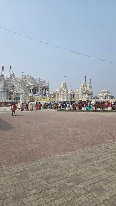 This image captures a bustling religious complex, likely a Jain temple site in Madhuban, India, under a clear, bright blue sky during daytime. A long procession of intricately carved white temples, characterized by their multiple spires (shikharas) and detailed architecture, forms the primary subject on the left side of the frame. These structures extend into the distance, suggesting a large complex.

Numerous people are visible throughout the scene, gathered around the temples and along the wide, paved pathway that occupies the right and central foreground. Many individuals appear to be devotees or visitors, dressed in traditional Indian attire, with women in colorful saris being particularly noticeable. They are seen walking, standing, and congregating, indicating active visitation and possibly religious observance.

The ground is a well-maintained paved area, distinctively split into two sections: a reddish-brown part closer to the viewer and a lighter gray section with a textured, possibly interlocked paver pattern. A faint, dotted line or trail is visible against the sky in the upper left. The clear sky and bright illumination suggest sunny weather. The overall atmosphere conveys devotion and community at a significant pilgrimage location. No visible text is present in the image.