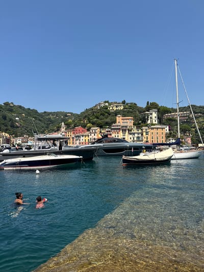 The image depicts a sunny day in the harbor of Portofino, Italy. In the foreground, two individuals are swimming in the clear blue water. A woman in a dark swimsuit and sunglasses is on the left, and a child wearing a red and white inflatable toy is to her right. The water is a bright turquoise, with sunlight creating dappled patterns on the seabed visible in the lower right corner.
Several boats are anchored in the harbor. Closest to the swimmers is a sleek, white speed boat with blue and red trim. Behind it, a larger gray motor yacht is docked. Further out, a classic sailboat with a white hull and tall mast is moored. Another smaller boat, covered with a beige tarp, is situated between the motor yacht and the sailboat.
The background is dominated by the picturesque hillside of Portofino, dotted with colorful buildings. The architecture features pastel facades in shades of yellow, pink, orange, and green, set against a backdrop of lush green trees. The sky is a clear, vibrant blue. The scene captures a moment of leisure and the iconic beauty of the Italian Riviera.