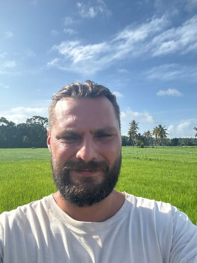 The image is a selfie taken outdoors in Thihagoda, Sri Lanka. The subject is a man with a beard and short hair, wearing a white t-shirt. He is positioned in the foreground, with his face partially visible. Behind him, a vibrant green rice paddy stretches out, bordered by a line of palm trees and lush vegetation. The sky is a clear blue, dotted with wispy white clouds. The bright sunlight suggests it is daytime, likely late morning or early afternoon. The overall scene evokes a sense of natural beauty and tranquility. There is no visible text in the image.